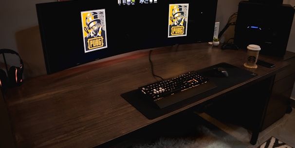 A gaming setup featuring a large curved monitor displaying PUBG logos on the screens. A keyboard and mouse are positioned on a large desk pad. A pair of headphones rests on the desk alongside a mobile phone. A drink in a Starbucks cup is placed to the right, while a desktop tower is seen in the background on the right side.