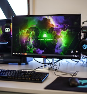 A gaming setup featuring a central monitor with a vibrant display showing 'The Spirit of Gaming'. On either side, there are gaming PCs with illuminated components and transparent cases. Headsets hang from the side of the monitor. The desk also has a black keyboard and mouse, and a branded mousepad.