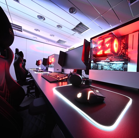 A row of high-end gaming computers on sleek desks, featuring RGB lighting and gaming accessories. The computers are housed in transparent cases with visible components, emitting a red glow. Comfortable gaming chairs with ergonomic design are positioned next to the desks, and the room's ceiling is modern with recessed lighting.